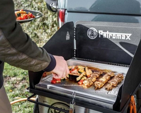 Persoana ce prajeste carne si legume pe placa de inox a gratarului Petromax